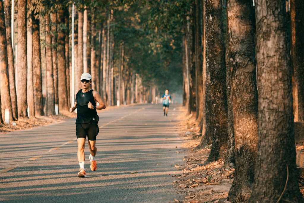 Two runners exercising on a serene forest road lined with tall trees, under warm daylight.